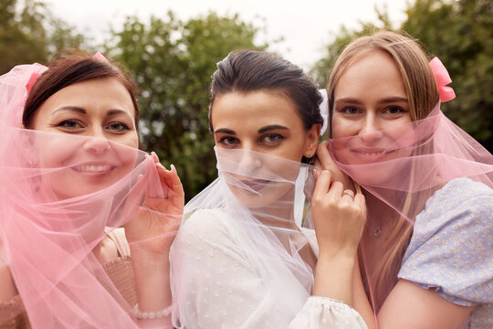 A happy charming bride in a white dress with a white veil and two bridesmaids in a blue and beige dress with pink veils smile and playfully cover their faces with veils. Cheerful bachelorette party.