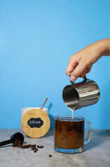 Pouring milk into black coffee in a glass, stevia in the background with blue background