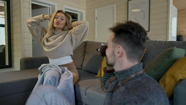Joyful woman posing sitting on couch as man taking photos with camera in living room. Cheerful relaxed happy Caucasian couple having fun on weekend enjoying leisure indoors