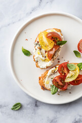 Bruschetta sandwiches with tomatoes, cream cheese, olive oil and basil on a plate on white marble background, top view, copy space. Traditional italian antipasti