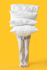Young woman in pajamas and with pillows on yellow background
