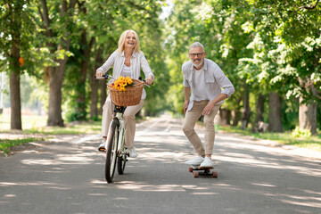 Modern Pensioners. Happy Senior Couple Riding Bike And Skateboard In Park