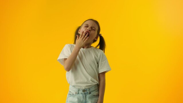 Young girl looks tired, feeling bored, and yawns widely, covering her mouth with a hand on a yellow background. Concept of the lack of interesting activities for the child