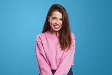 Fototapeta premium Happy young Hispanic woman in pink sweater standing with holding hands together and looking at camera happily, over blue background