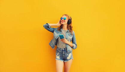 Portrait of stylish modern happy young woman listening to music in headphones with phone wearing jacket, sunglasses on yellow background
