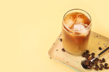 Glass of ice coffee with beans, spoon and plate on yellow background