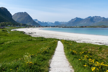 une sentier de bois blanc qui mène à une plage de sable blanc avec une mer bleue et des montagnes en fond