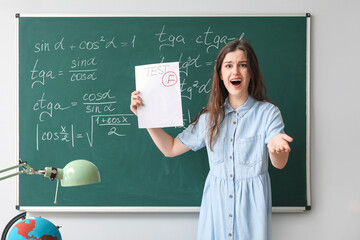 Displeased female teacher with checked test sheet near blackboard in classroom