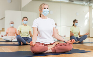 Group of people in protective mask sitting in lotus position practicing meditation in yoga class