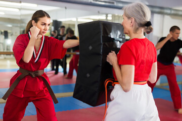 Fototapeta Young caucasian woman exercising jabs on punching pad in senior woman's hands during karate training. obraz