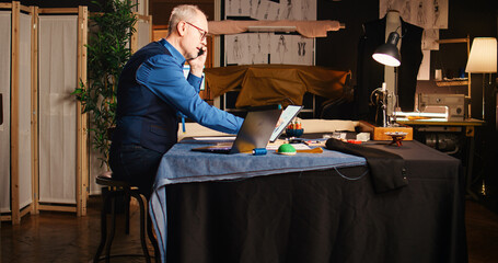 Old designer calling customer in atelier, discussing about measurements and fabric on phone call. Senior tailor working on custom made clothing line to create tailored items. Handheld shot.