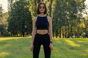 Young sport female in in black tight sportswear before fitness training session at the park. Healthy young woman warming up outdoors. Fitness outdor. hot summer day