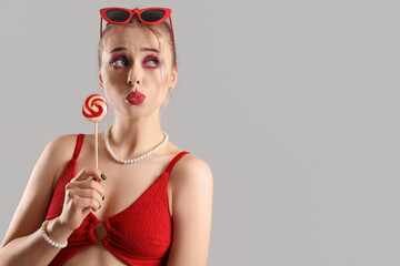 Thoughtful young woman in swimsuit with lollipop on grey background, closeup
