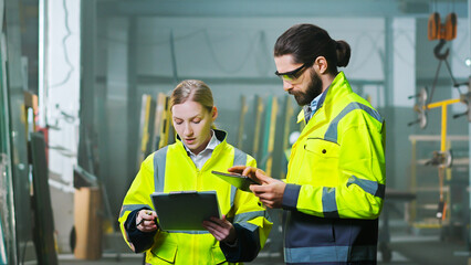 Obraz premium Caucasian man and woman colleagues at manufactory talking and discussing work with tablet device and documents or plan. Production plant. Indoors. Male and female co-workers discussion.