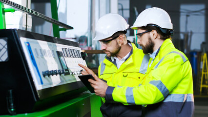 Caucasian male worker of plant in helmet and yellow uniform working at controlling point and colleague helping. Indoors. Men in hardhats at controller in factory. Co-worker with tablet asking for help