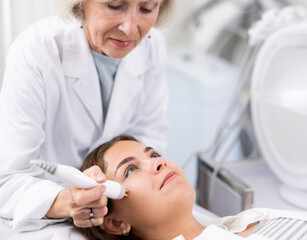 Young woman receiving non-surgical rejuvenation procedure from senior cosmetologist, RF face lifting with use of electric device equipment in spa center