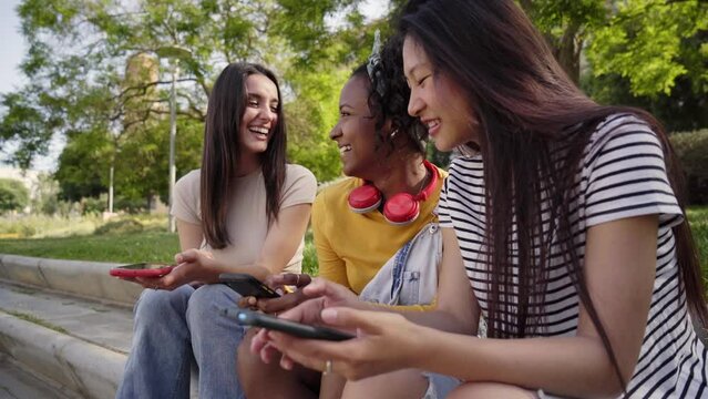 Three smiling young women using cell phone outdoors. Group of happy friends checking fun social networks on mobile. Cheerful classmates gossip on dating apps together. People and new technologies.