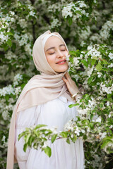 A young Muslim woman in a white dress in front of a flowering tree. Medium plan. Blooming apple tree. Pretty modest Muslim woman. European Muslim. Tenderness. Beauty.