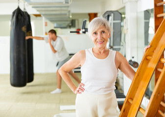 Senior caucasian woman standing at gym ladder, smiling and looking at camera.