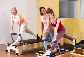 Young woman practicing pilates system on reformer supervised by qualified latin american female trainer..