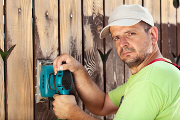 Male worker polishing old wooden fence with power tool - a vibrating sander