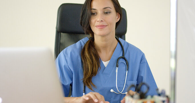 Beautiful female doctor with stethoscope grinning while typing on laptop computer at desk with black leather chair in office