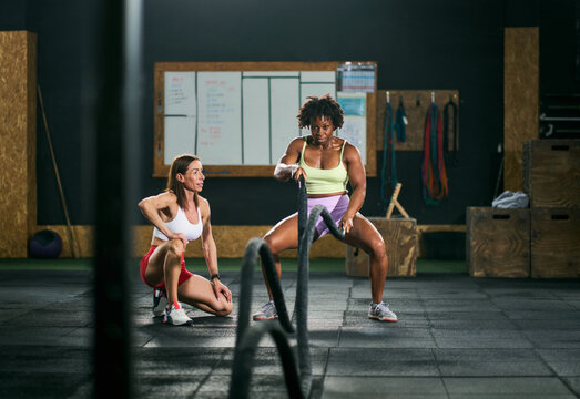Friend supporting black sportswoman doing exercise with battle ropes