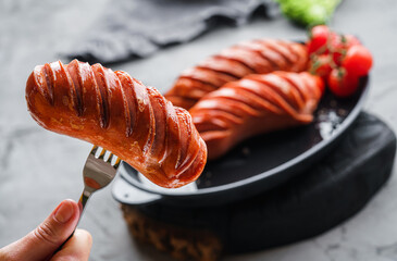 Grilled smoked sausages in pan in dark wooden background with tomatoes and spices. Hot sausage on a fork, meat food, selective focus - obrazy, fototapety, plakaty