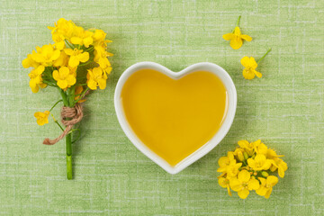 Rapeseed oil in heart shaped bowl and flowers on green background, top view.