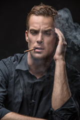 Smoking man in dark shirt on the black background in the studio. He holds the left hand near his head and looks down. Smoke swirls around him. Vertical low-key photo.
