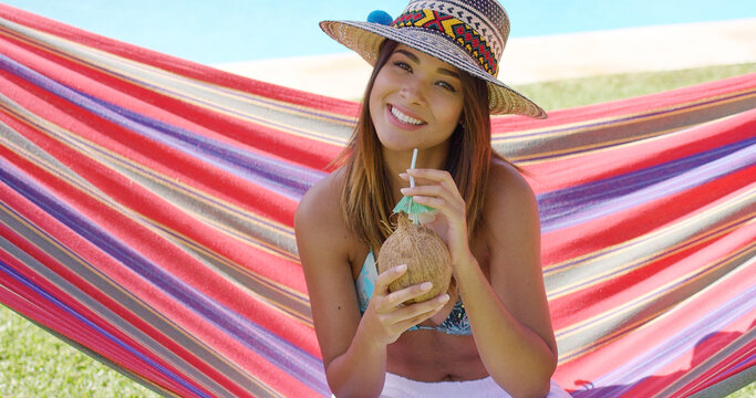 Cute young woman in hat and bikini top with white pants holding coconut drink while sitting in hammock