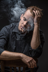 Stylish guy in dark shirt sits on a chair on the black background in the studio. He smokes a cigarette with closed eyes. Left hand is on the head, right hand is on the chair. Vertical low-key photo.
