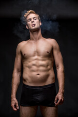 Fototapeta na wymiar Macho with muscular body in black underpants stands on the black background in the studio. He smoke a cigarette and a smoke swirls around him. Vertical low-key photo.