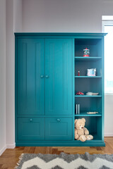 Kid's turquoise bookcase with toys, books, notebooks, pencils and a big teddy bear. On the floor there is a parquet with a gray carpet. Walls are white. Indoors. Vertical.