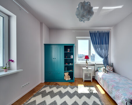 Room for kid with light walls and a parquet with carpet on the floor. There is a bed with colorful veil and pillows and a toy, turquoise bookcase with toys and books, nightstand with a lamp.