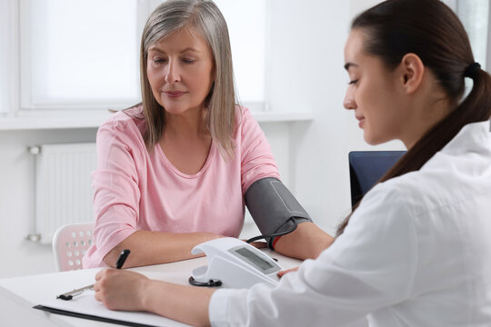 Doctor measuring blood pressure of woman and writing it down into clipboard at table indoors