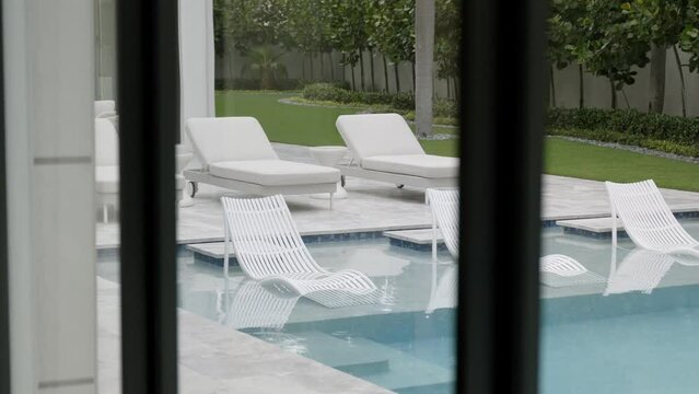 View from window on lounge area near inground swimming pool with six white deck chairs. Interior design of splendid backyard with large patio for rest