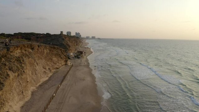 Aerial Shot Of People At Beach Near Hill And City Against Sky, Drone Flying Forward Over Sea At Sunset - Herzeliya, Israel