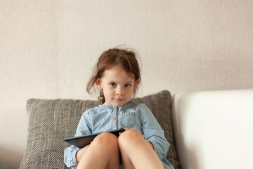 Little girl sitting on the sofa and holding a tablet in her hands