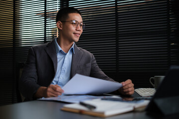 Asian businessman analyzing financial reports and using laptop at office.