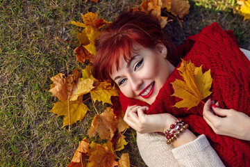 Beautiful young woman with red hair holding yellow maple leaf lying on the ground. Outdoor shot. Fall. Copy space.