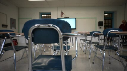 Teacher working in background while camera moves across the backs of empty school desks and chairs with smart board and US flag.