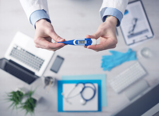 Doctor measuring temperature and holding a digital thermometer, desktop with medical equipment and computer on background, top view
