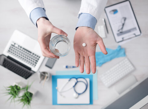 Doctor giving a pill and holding a glass of water, desktop with medical equipment and computer on background, hands close up top view