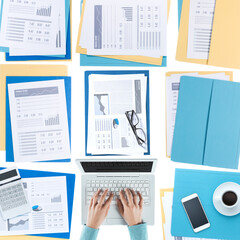 Business woman working at desktop on a laptop with financial reports, paperwork and files, top view, unrecognizable person
