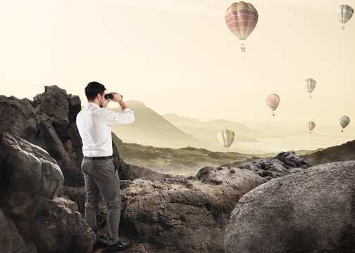 Businessman leaning on a rock watching hot-air balloons in the sky with binoculars