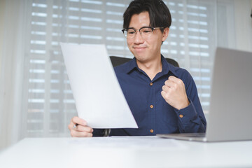 Emotional happy Asian businessman in business casual long shirt holds contract paperwork when was accepted, approved agreement contract. Young adult business man smiles when contract agreement passed - 622126333