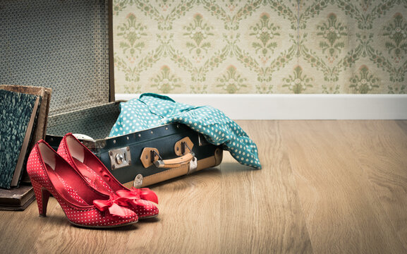 Open vintage suitcase with red shoes and dotted clothing, vintage wallpaper on background.
