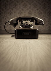 Black 1950s style phone on hardwood floor and vintage wallpaper on background.