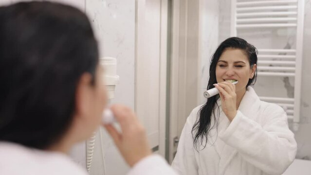 Cleaning teeth, Bathroom Beauty, Showering Woman. Striking adult woman cleans teeth following shower, observing reflection in bathroom mirror, adorned in white, satin bathrobe
